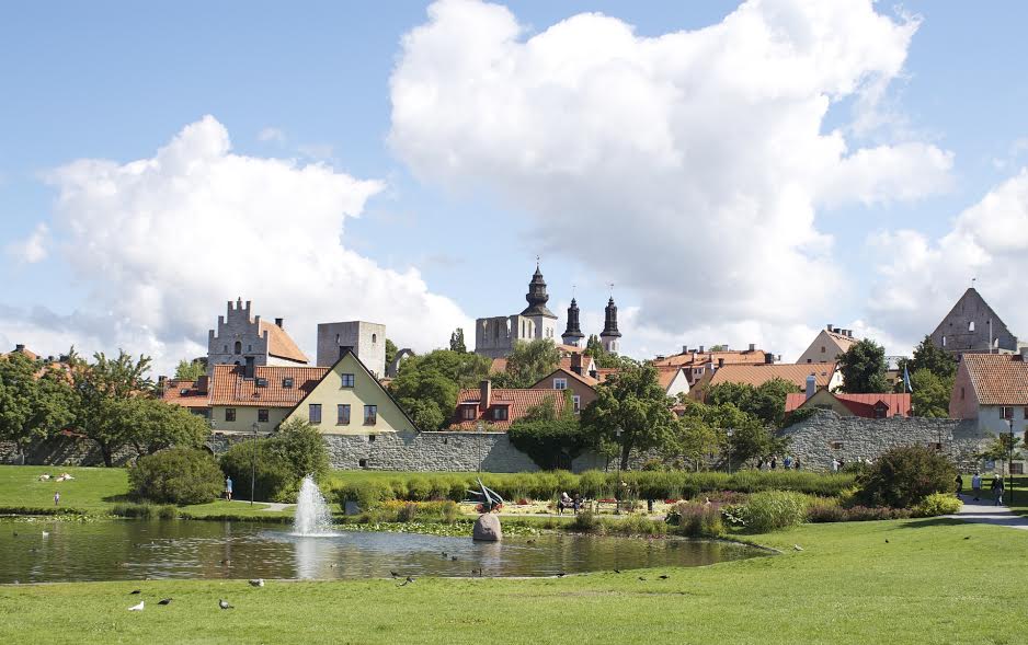 Staden Visby med hus och kyrktorn. Framför staden är en sjö med en liten fontän i.