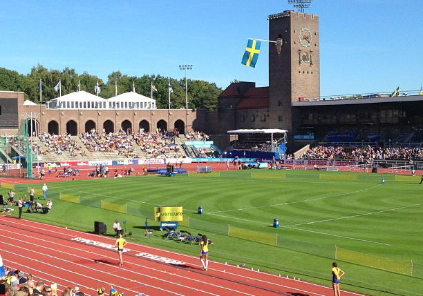 En stadio syns i solsken. På löparbanorna står några svenska idrottare.
