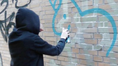 En person med mörk tröja och luvan uppdragen står vänd mot en vägg. Vi ser inte ansiktet. Personen har en sprejburk i handen och sprejar på en tegelvägg.