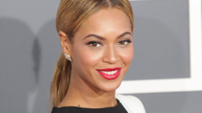 Beyonce står framför en grå vägg. Hon har blonderat hår i en mittbena och hästsvans. Hon har klarrött läppstift och svarta kläder.