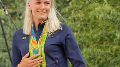 Sara Sjöström. Bakom henne syns massa gröna löv. Hon har långt ljust hår och en blå jacka. Runt halsen hänger flera medaljer i band.