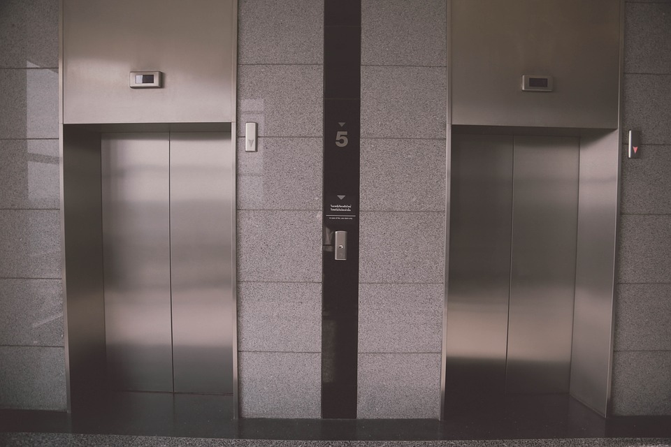 Bilden föreställer två hissdörrar, sida vid sida. Dörrarna är gjorda i ett silvrigt metallmaterial. Runt om dem är det grått kakel.