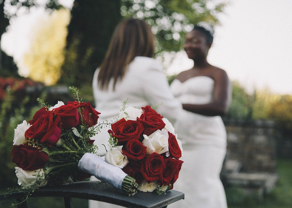 Två kvinnor som ska gifta sig står och håller händer med två rosbuketter framför dem.