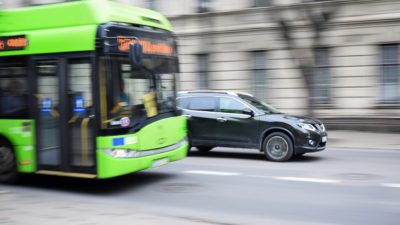 En grön buss åker på en väg. Bredvid den åker en svart bil. Bilden är lite suddig som om den har tagits i farten.