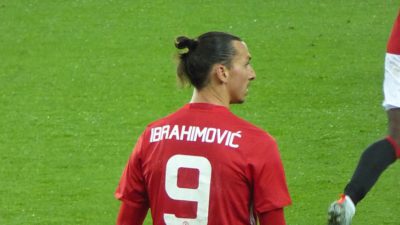 Zlatan står på en fotbollsplan med ryggen mot oss. Han tittar åt sidan och har en röd fotbollströja på sig. På ryggen står det Ibrahimovic och siffran 9 i vitt.