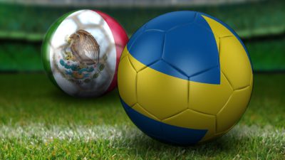 Två fotbollar ligger på en fotbollsplan. Den närmste bollen är helt blå med ett gult kors hela vägen runt. Den andra bollen är grön, vit och rödrandig. På det vita fältet syns en stor örn som sotter på en kaktus.