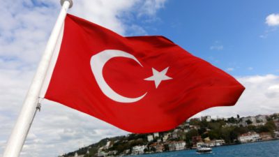 Turkiets flagga viftar i vinden över vatten. Det ser ut som att den sitter längst bak på en båt. Flaggan är röd med en vit halvmåne och en vit stjärna.