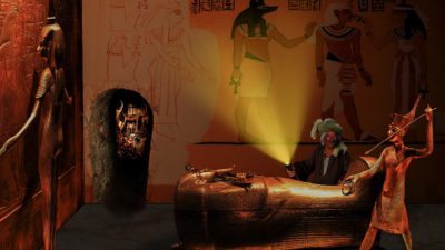 En egyptisk gravkammare med en kista som är formad som en person. Runt kistan står det statyer och bakom kistan står det en person med ficklampa och ser glad ut.