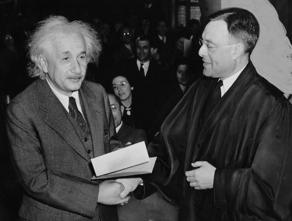 Einstein står och skakar hand med en person i lång svart kappa. Einsten har stort vitt yvigt hår och ser lite finulrig ut.