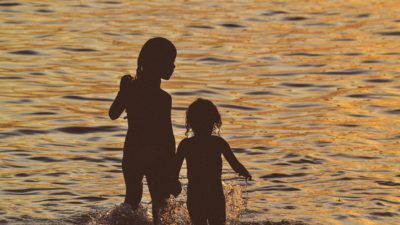 Två barn står med vatten upp till knäna i vattenbrynet. Det är kväll så det går inte att urskilja detaljer utan bara barnens siluetter.