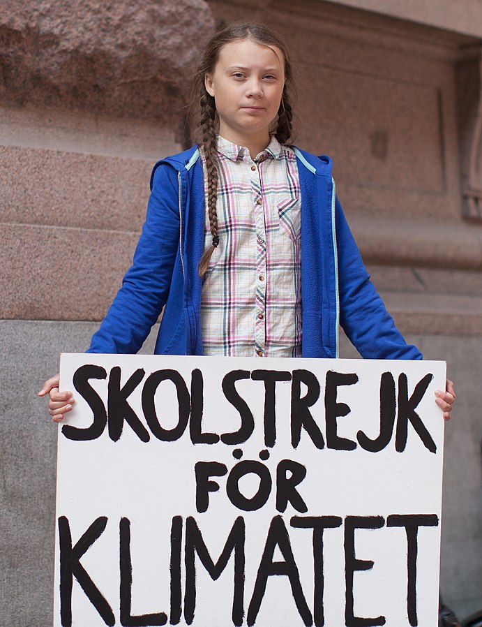 Greta Thunberg står i en rutig skjorta, en blå tjocktröja och håller i en skylt där det står Skolstrejk för klimatet