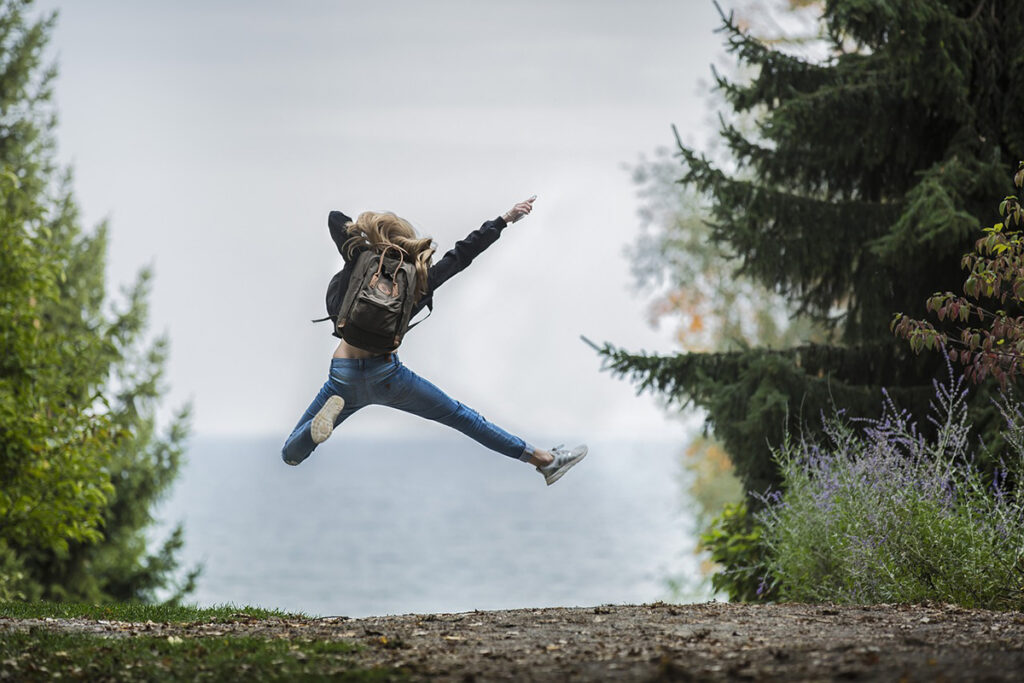 En person som hoppar högt upp i luften på en skogsväg med havet framför sig.